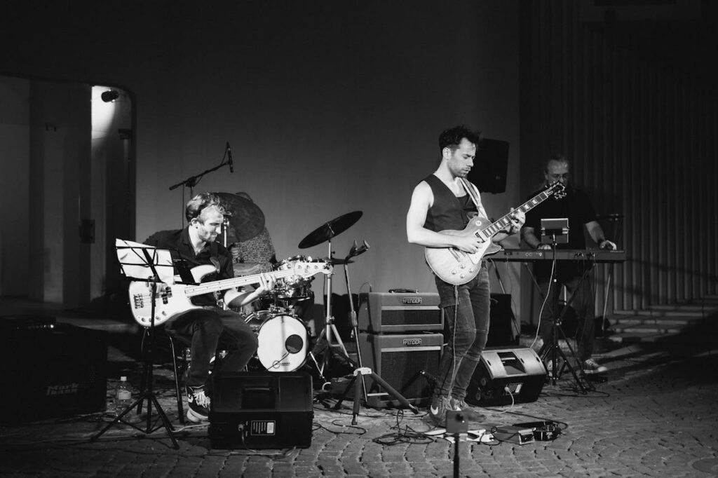 A dynamic black and white photo of a live jazz band performing with guitar, bass, and piano on stage.